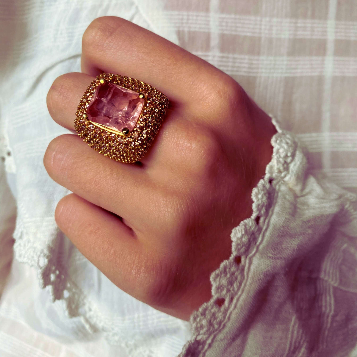 A person's hand wearing AVA, yellow gold plated statement ring with champagne cubic zirconia stones, featuring a large champagne pink doublette in the center.
