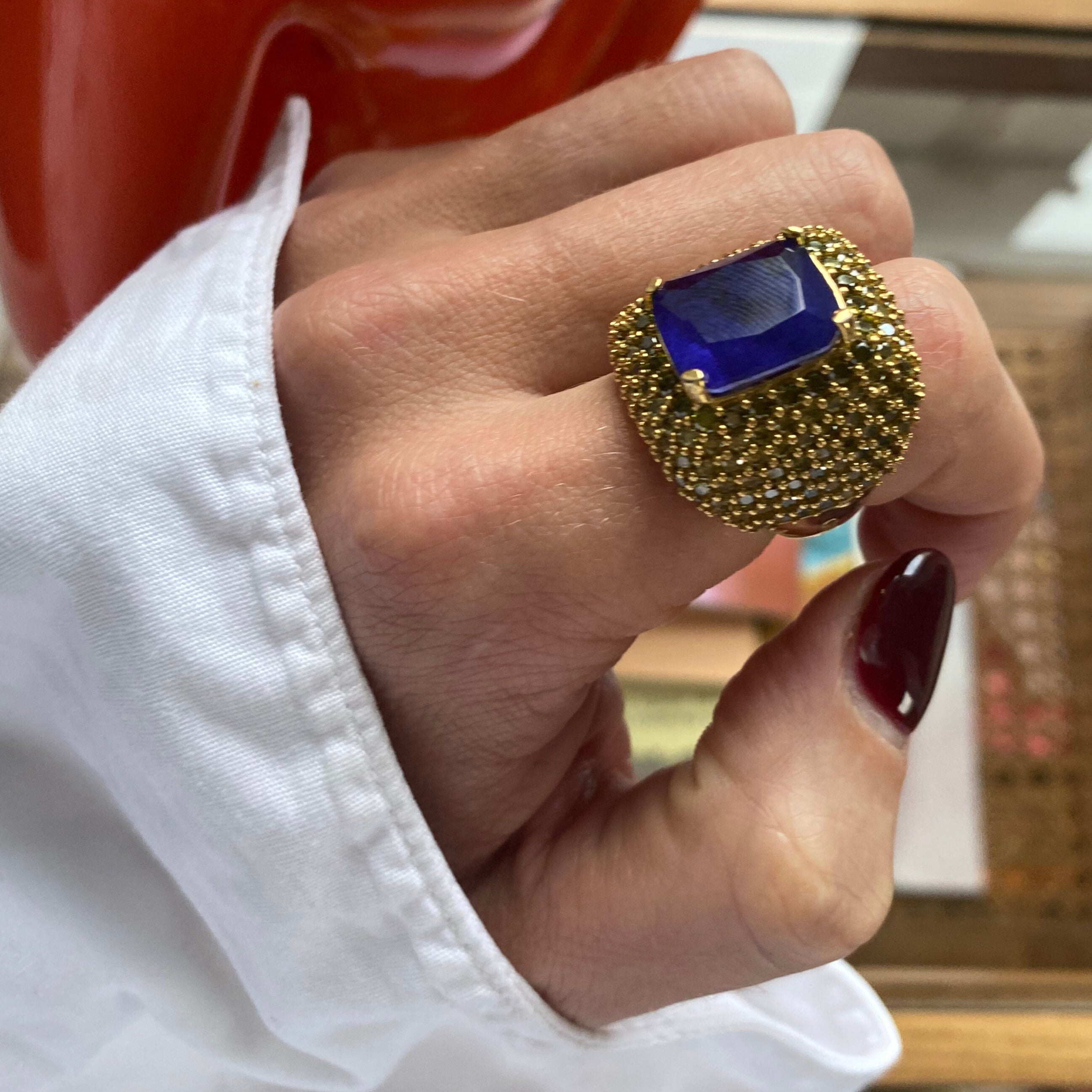 MALIKA blue cocktail ring angled view on hand. Showing yellow gold plated ring with peridot green cubic zirconia stones and a large deep blue doublette center stone.