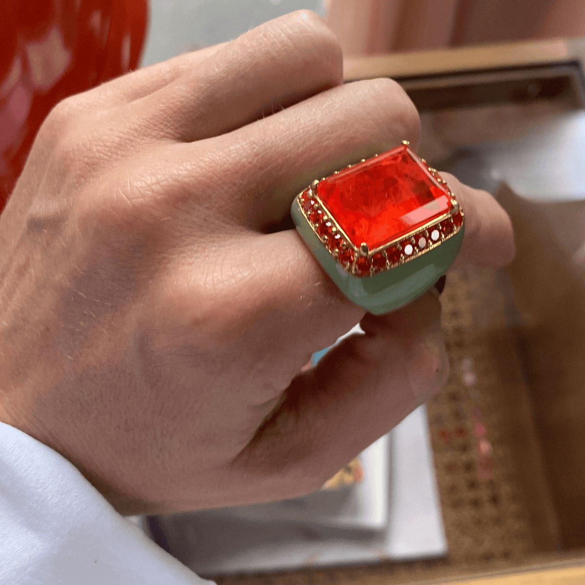 A person's hand displaying a yellow gold plated oversized statement ring with pastel green enamel and a large orange doublette surrounded with orange cubic zirconias - FREDDY by Giadan for Le.Paul Jewelgarden