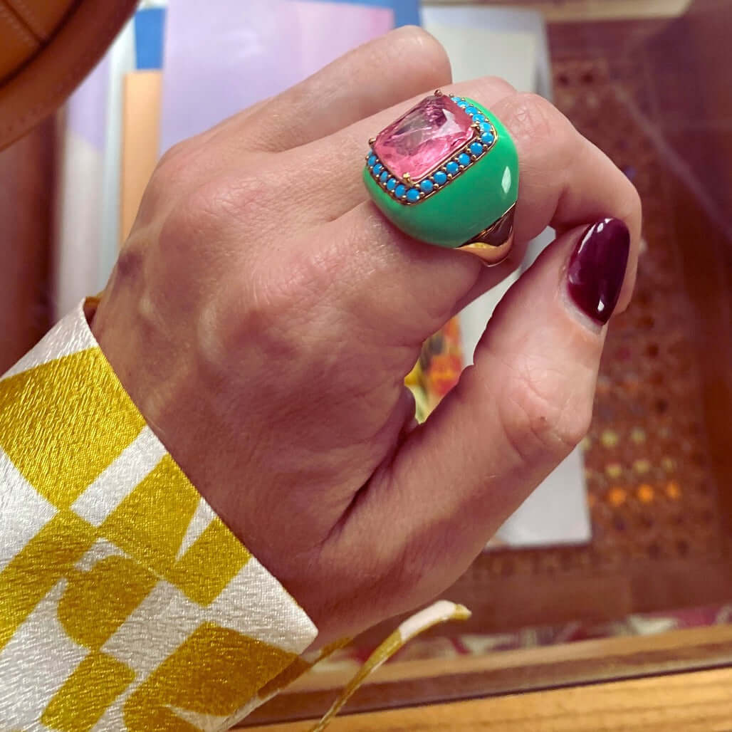 A hand wearing a yellow gold plated ring with mint green enamel and turquoise blue cubic zirconias surrounding a large pink doublette center stone.