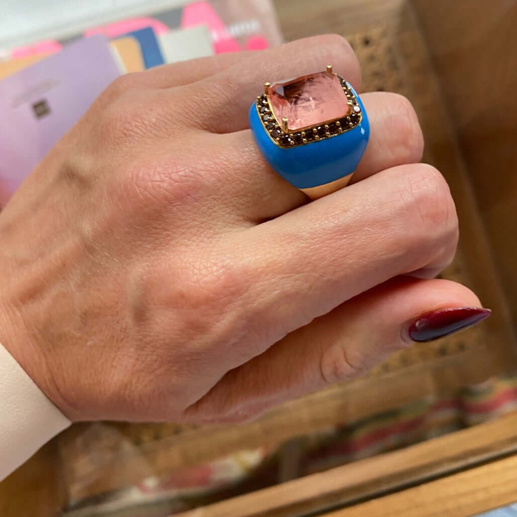 A hand wearing a blue enamel and yellow gold plated ring with a large pink champagne doublette surrounded by brown cubic zirconias.