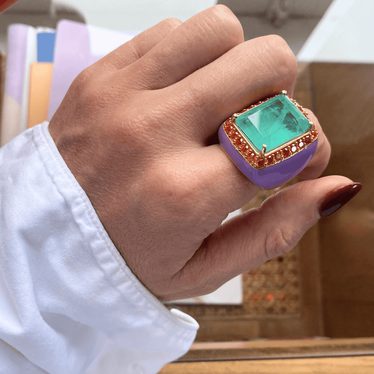 A person wearing a yellow gold plated ring with lilac enamel and aquamarine green doublette, surrounded by orange cubic zirconias - LOTTE by Giadan for Le.Paul Jewelgarden