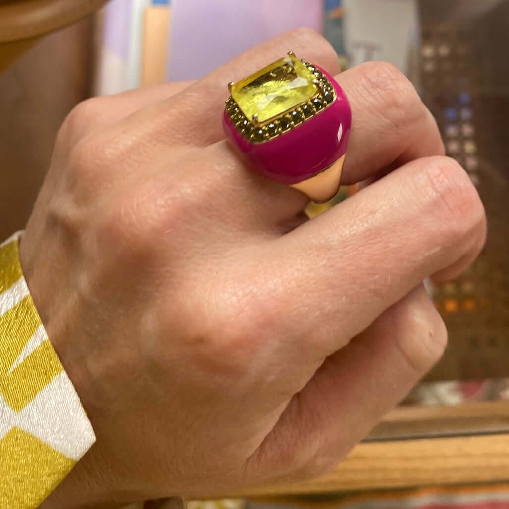A yellow gold plated ring with fuchsia pink enamel and a large yellow doublette surrounded by peridot green cubic zirconias, displayed on a person's finger.