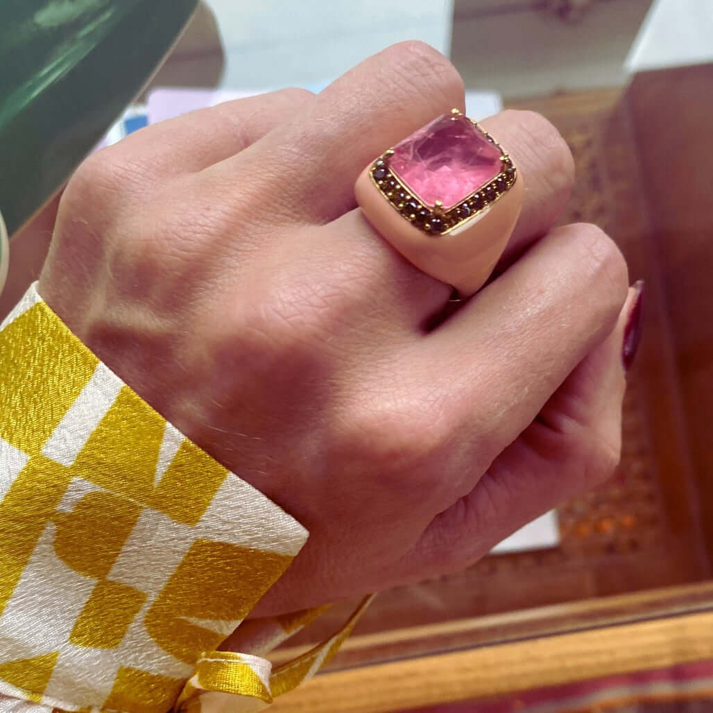 A person wearing a yellow gold plated ring with peach pink enamel and peridot green cubic zirconias and a pink doublette center stone.