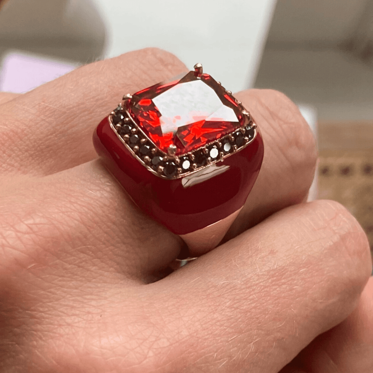 A person's hand displaying a rose gold plated ring with blood red enamel and a large rubellite red zirconia surrounded by smaller brown zirconias.