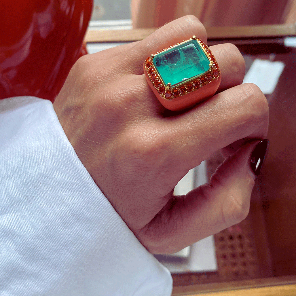 A person's hand wearing YASMINE ring - a yellow gold plated XL statement ring with salmon enamel surrounded by orange cubic zirconias, featuring a large aquamarine green doublette as the center stone. By Giadan for Le.Paul Jewelgarden