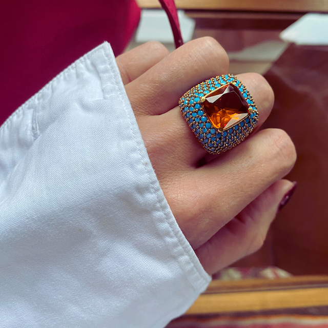 A close-up image of AFET yellow gold plated statement ring with a large central citrine yellow zirconia and turquoise blue zirconia stones on a person's hand.