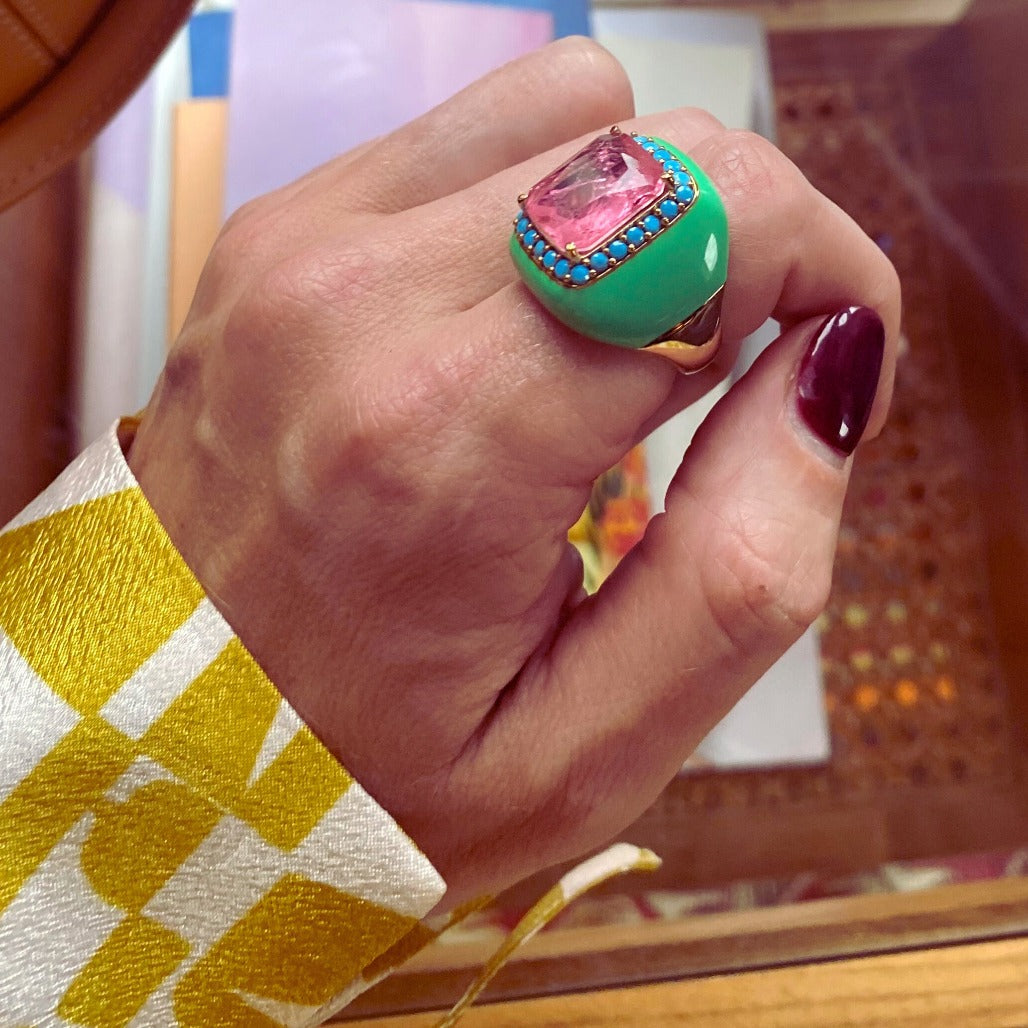 A hand wearing a yellow gold plated ring with mint green enamel and turquoise blue cubic zirconias surrounding a large pink doublette center stone.
